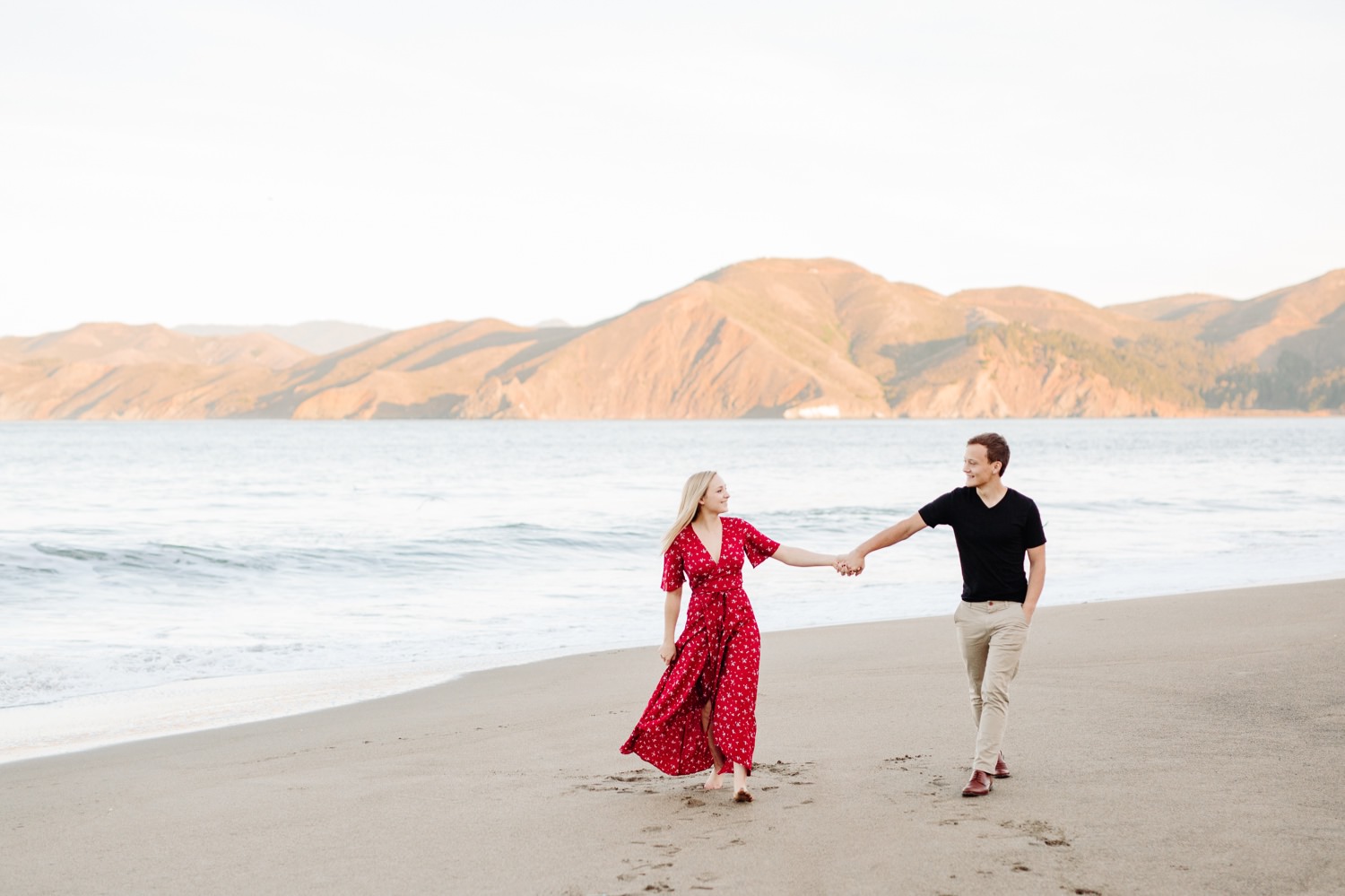 couple walking holding hands at engagement session at sunrise on bakers beach in san francisco
