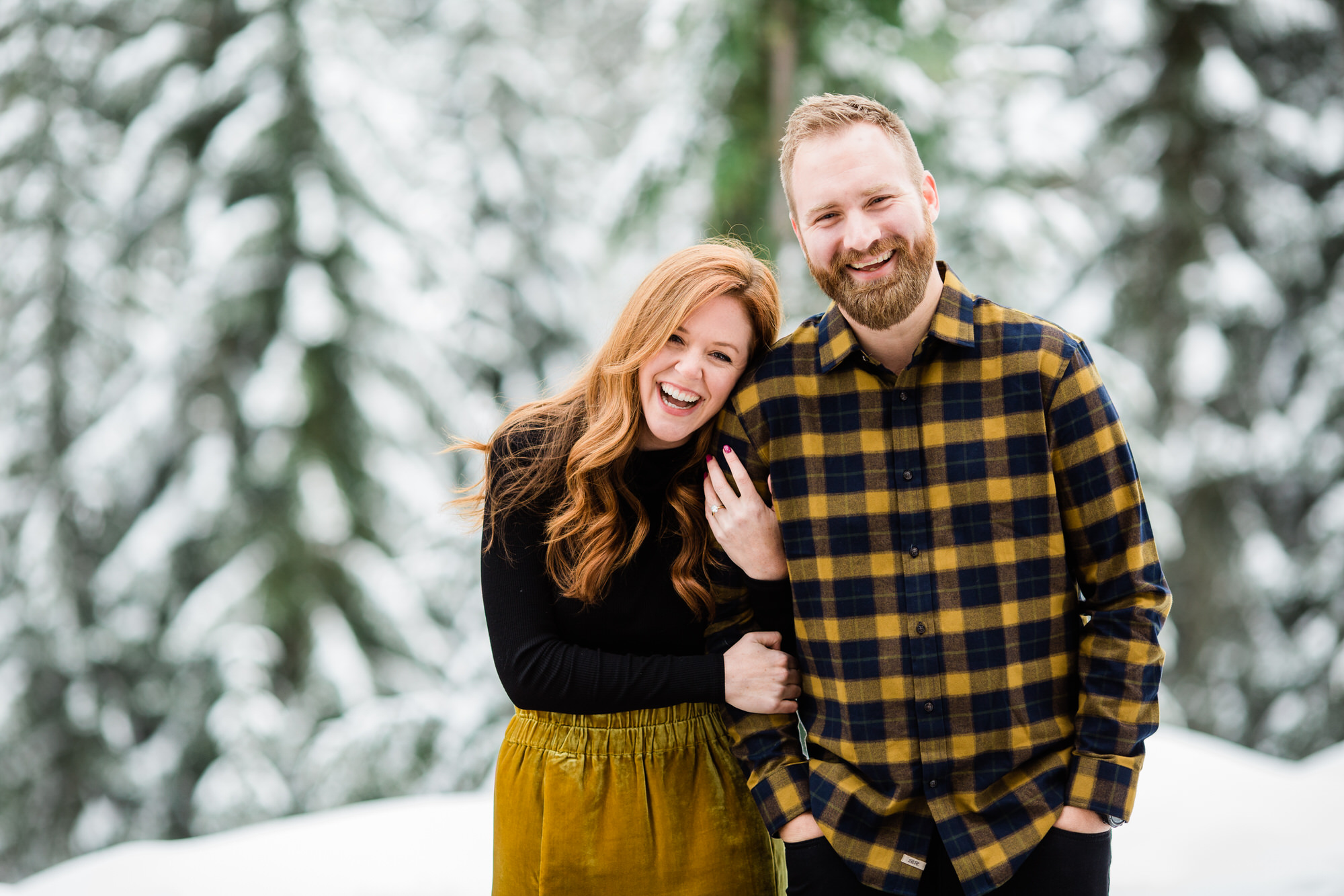 Couple leans on each other in front of snow-covered trees.
