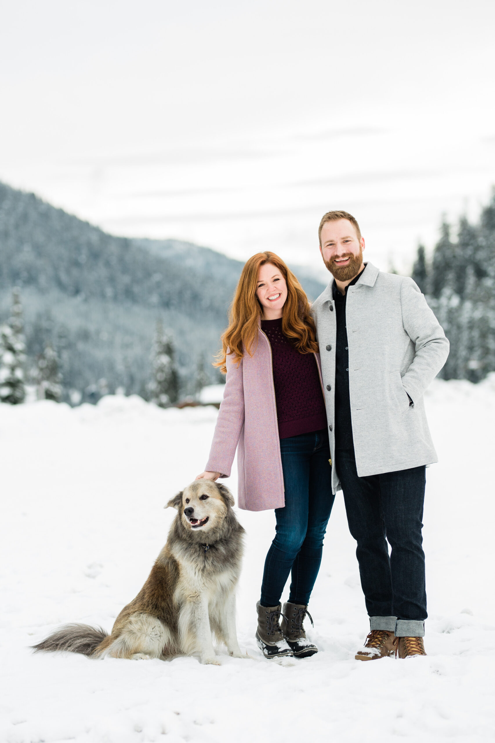 Couple stands together with their dog and poses for engagement photos in the snow.