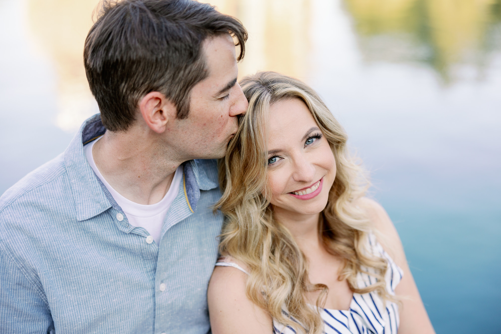 A couple cuddles together with a soft blue and green background of trees reflected in water at their engagement session at UW.
