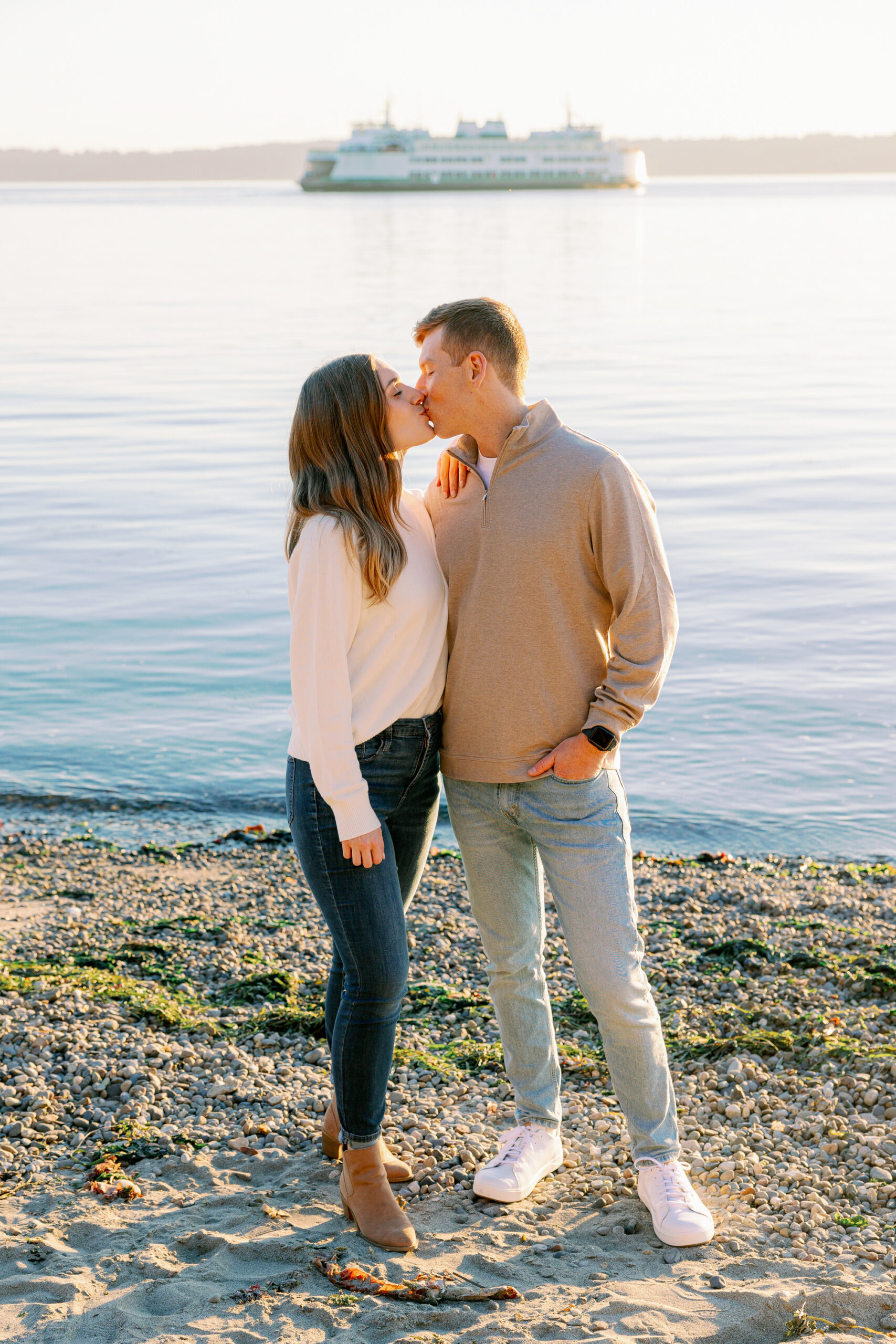A couple kisses during their engagement session with Puget Sound and a ferry in the background at Lincoln Park.