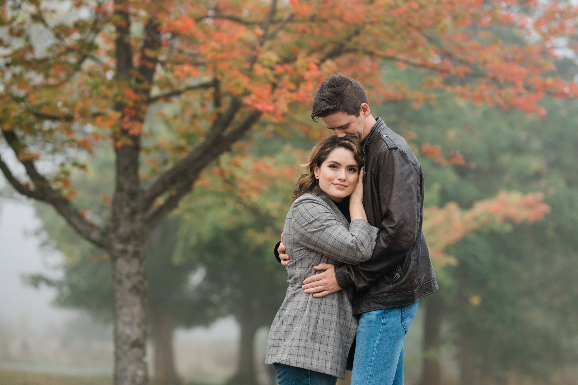Couple hugs in front of trees with fall colors on a foggy morning during their engagement session in Seattle.