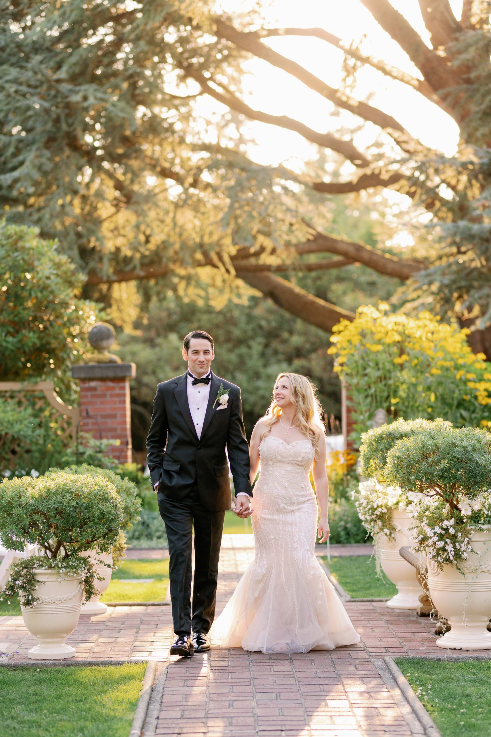 A bride and groom hold hands while walking through the garden at Lairmont Manor during sunset.