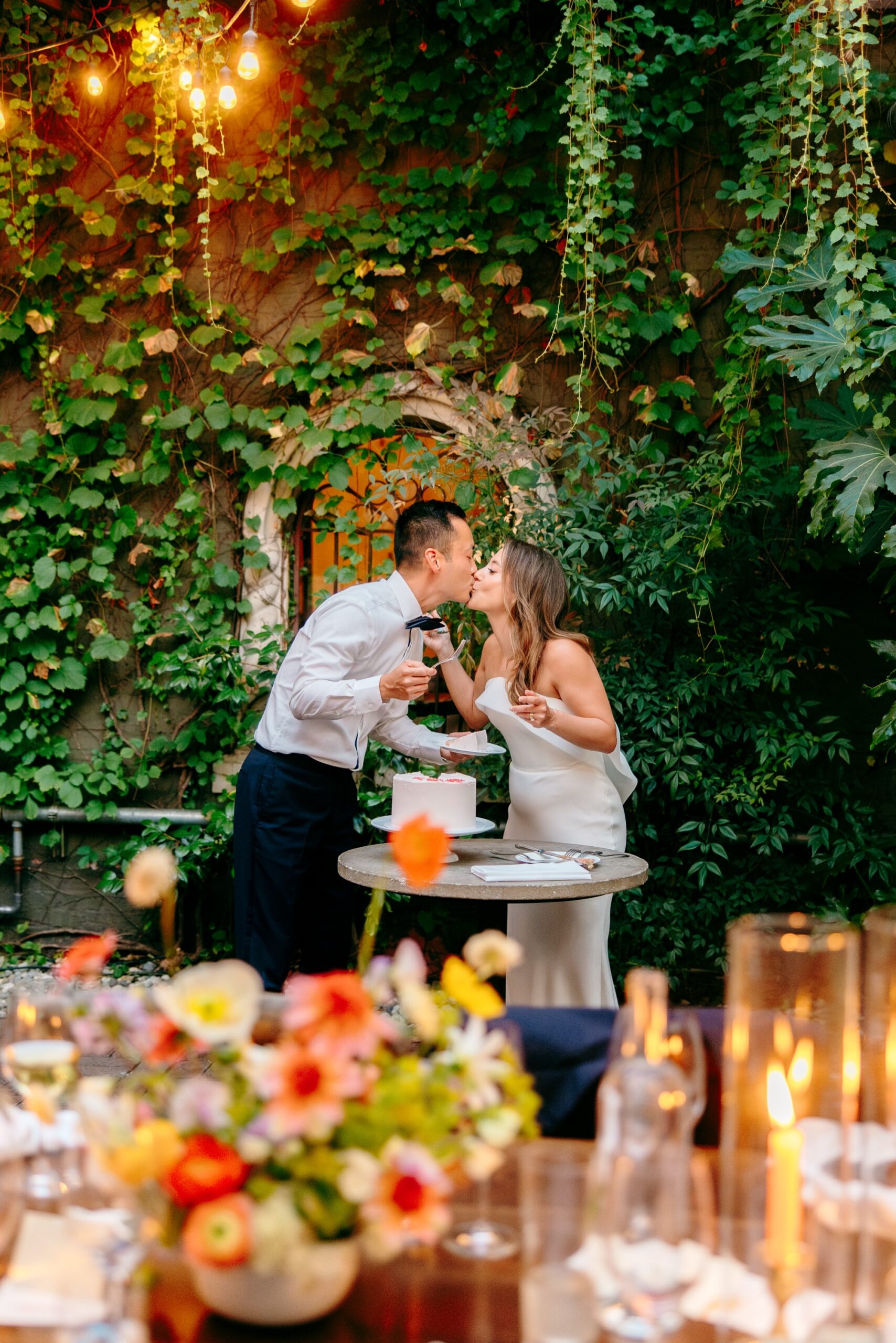 Couple kisses after cutting their wedding cake with the vine-covered Corson Building in the background