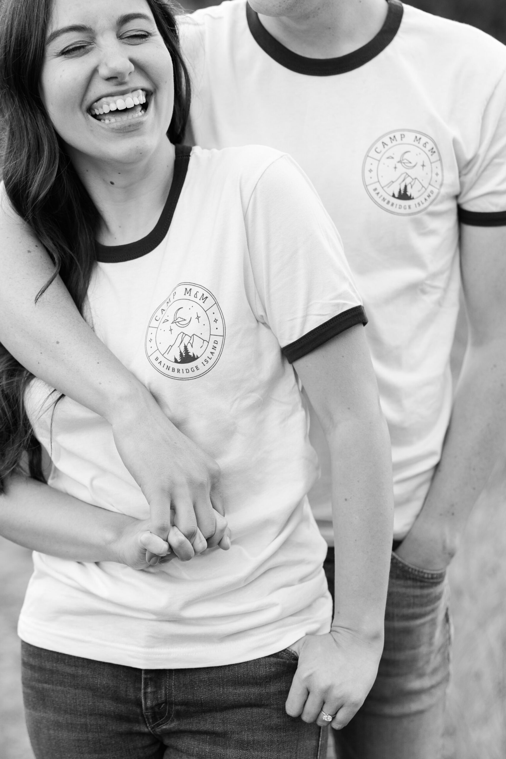 A black and white photo of a couple laughing and holding hands in matching "Camp M&M" shirts.
