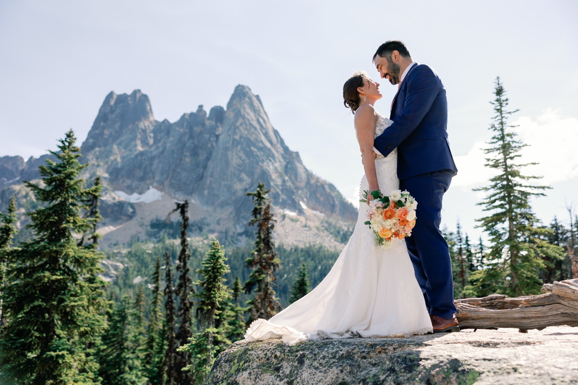 Bride and groom embracing on a mountain peak with a stunning backdrop of rugged mountains and pine trees.