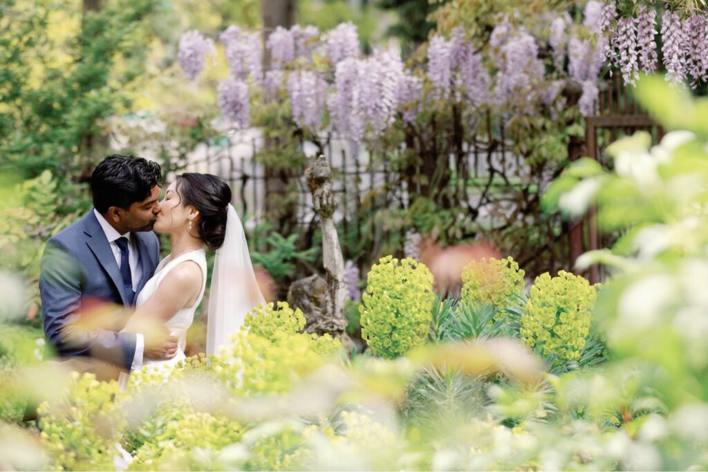 The bride and groom share a kiss surrounded by vibrant garden blooms and wisteria.