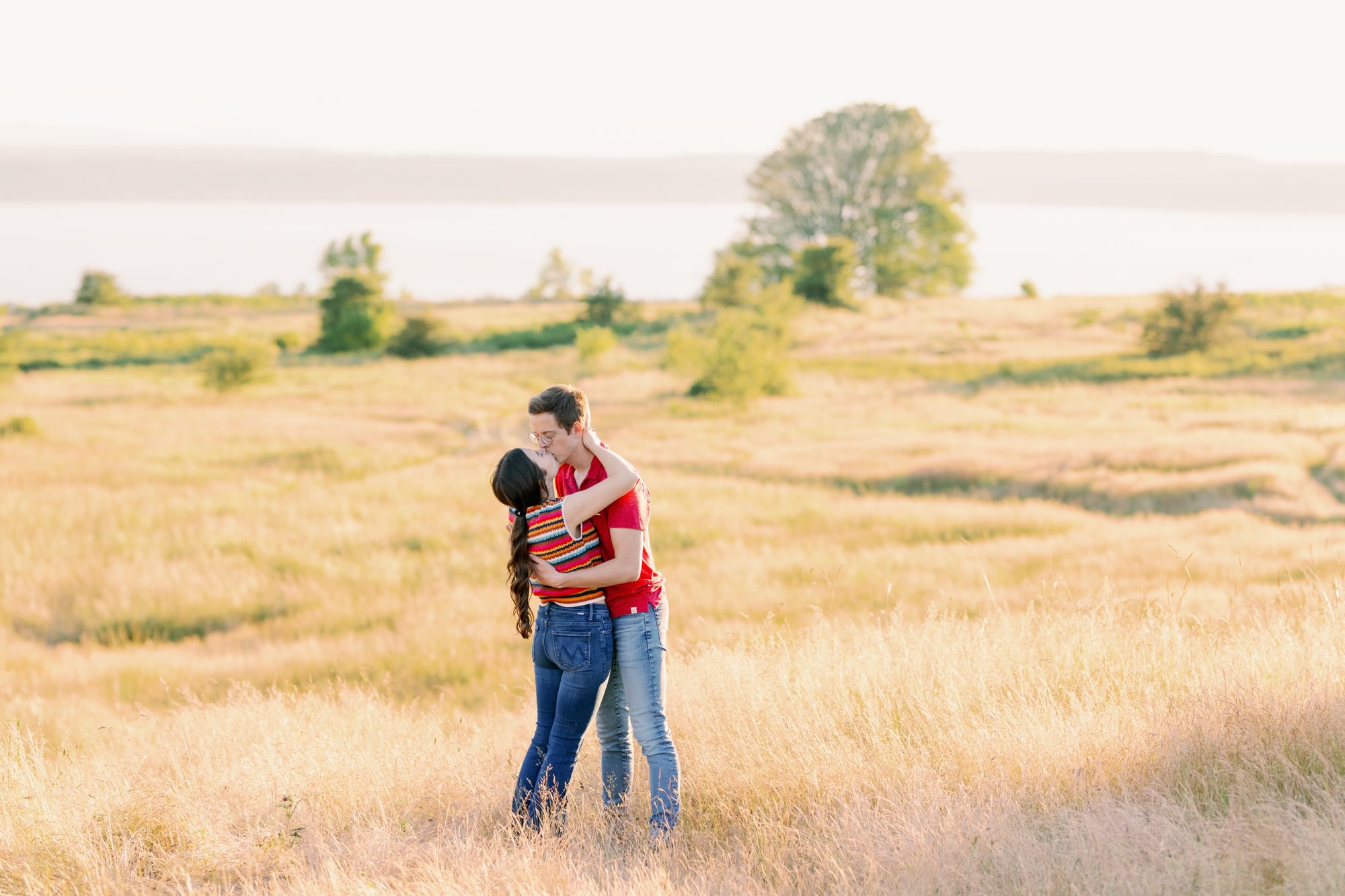 A couple sharing a kiss in a golden field with a scenic background of trees and water.