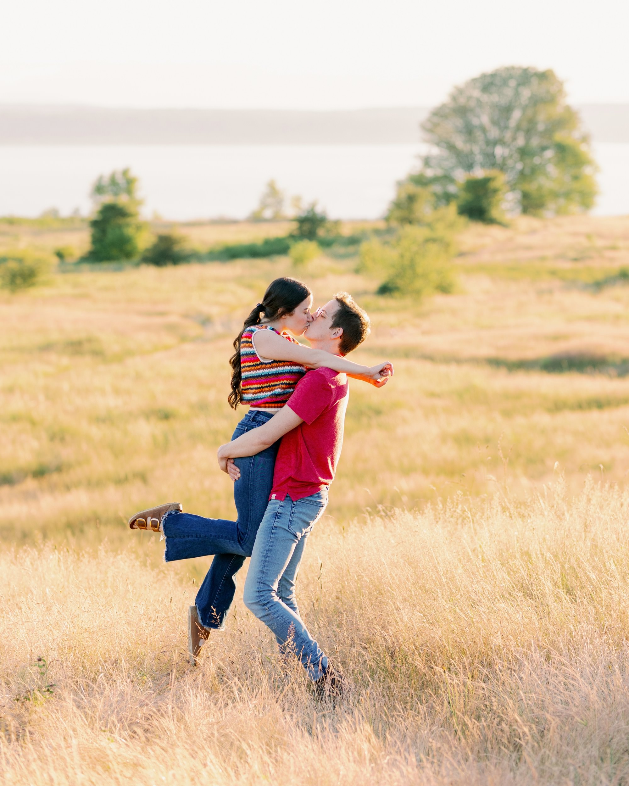 A man lifting his partner in a sunny field, both sharing a kiss and enjoying the sunset.
