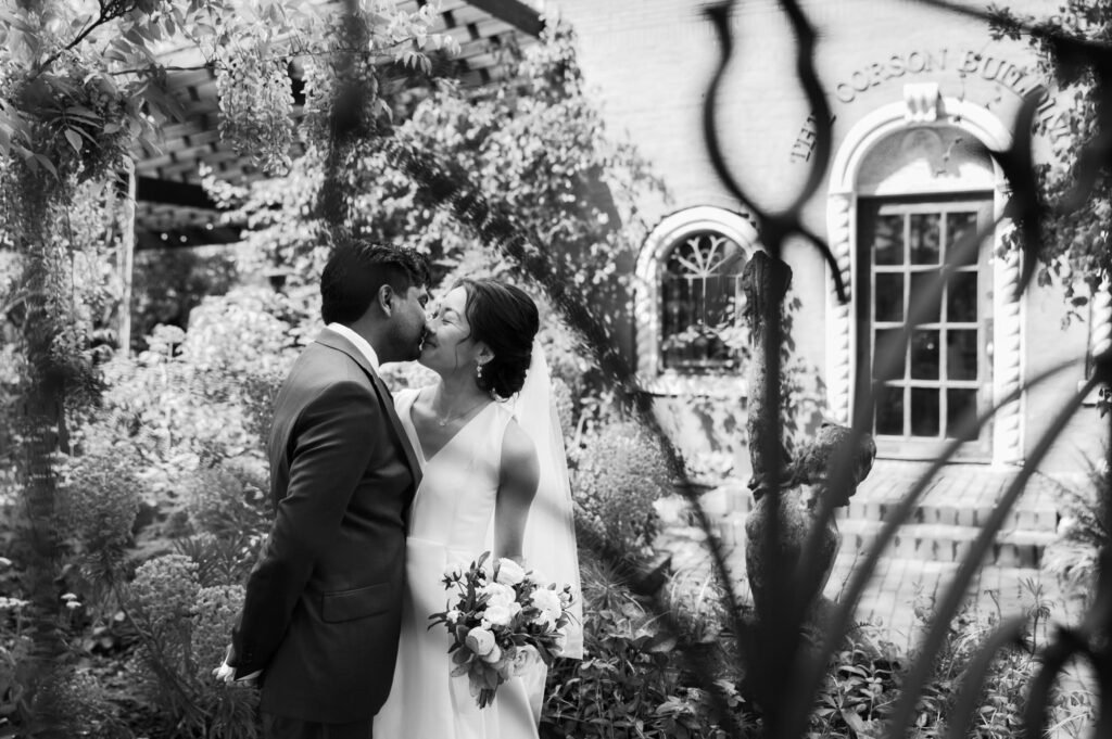 The bride and groom kiss in front of the Corson Building, surrounded by garden greenery.