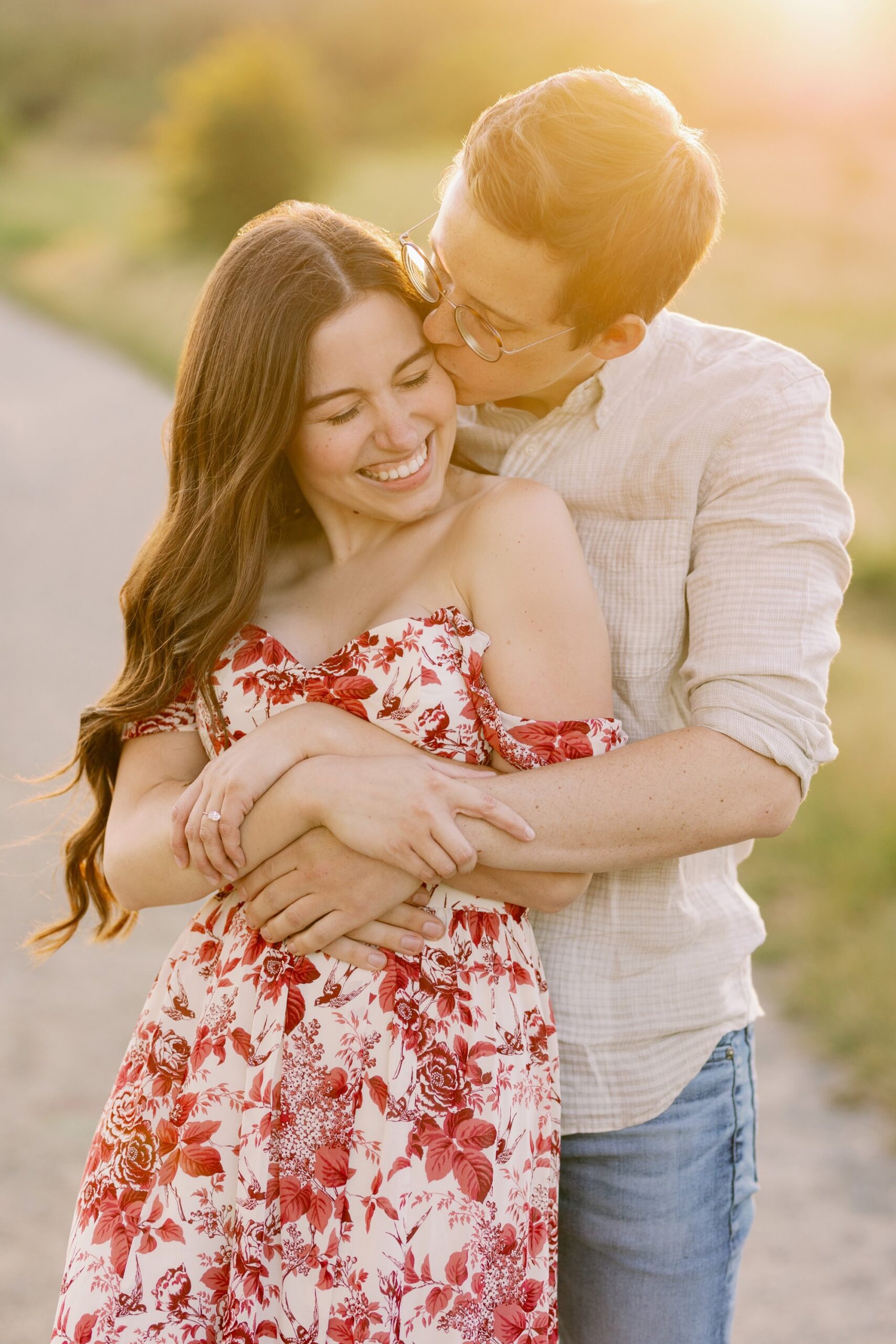 A man embracing his partner from behind and kissing her cheek, both smiling in the warm golden hour light.