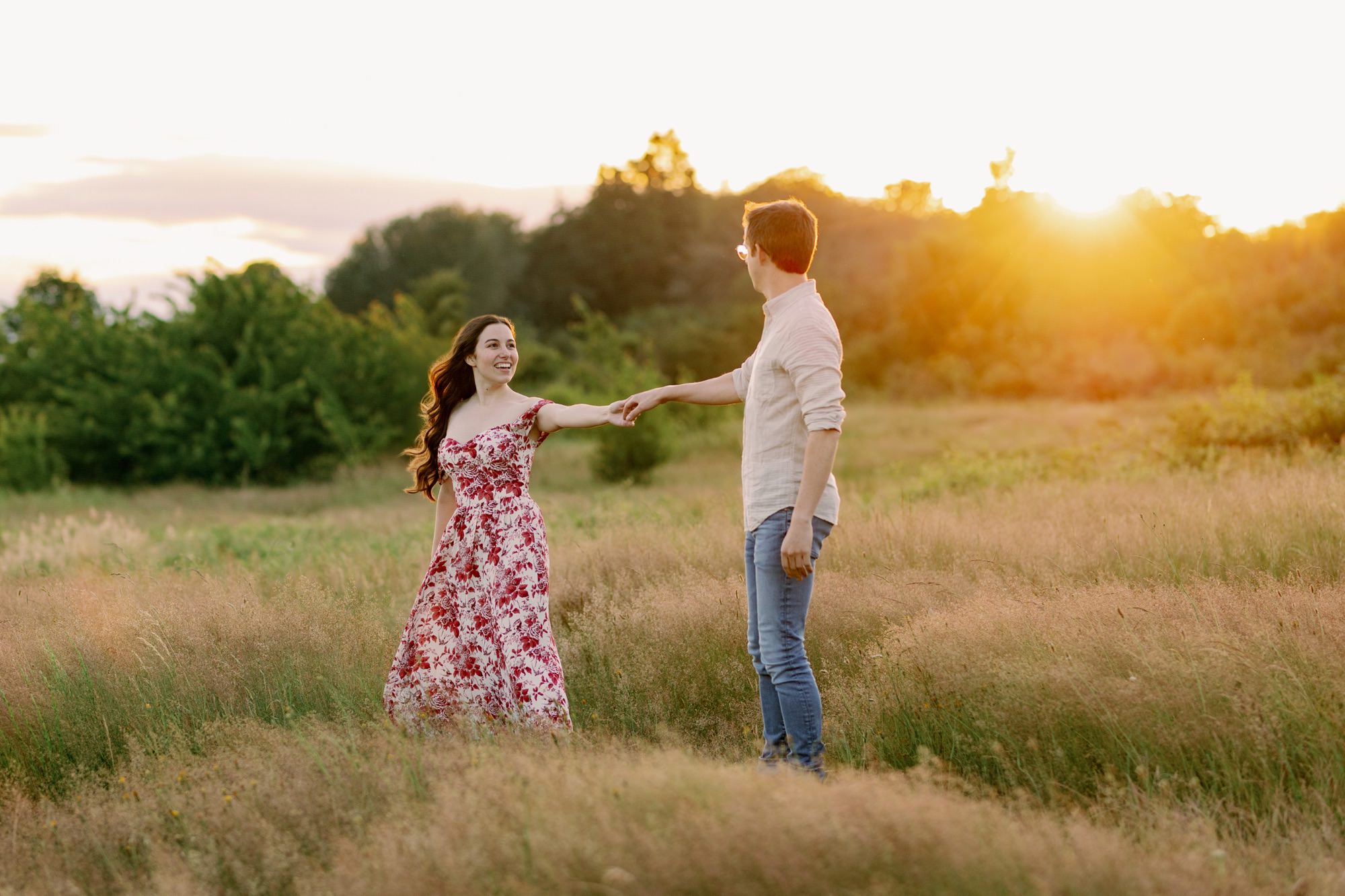 A couple holding hands and walking in a grassy field at sunset, with the woman in a floral dress and the man in a light shirt.