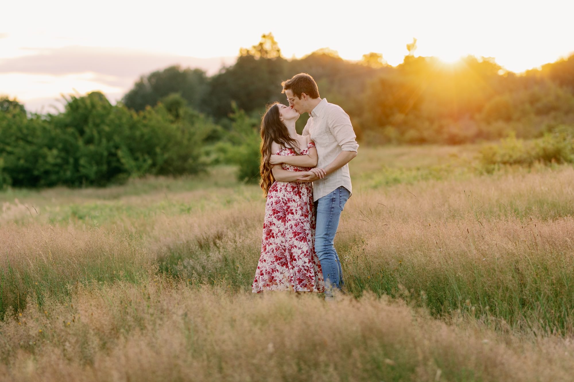 A couple sharing a kiss in a grassy field at sunset, with the woman in a floral dress and the man in a light shirt.
