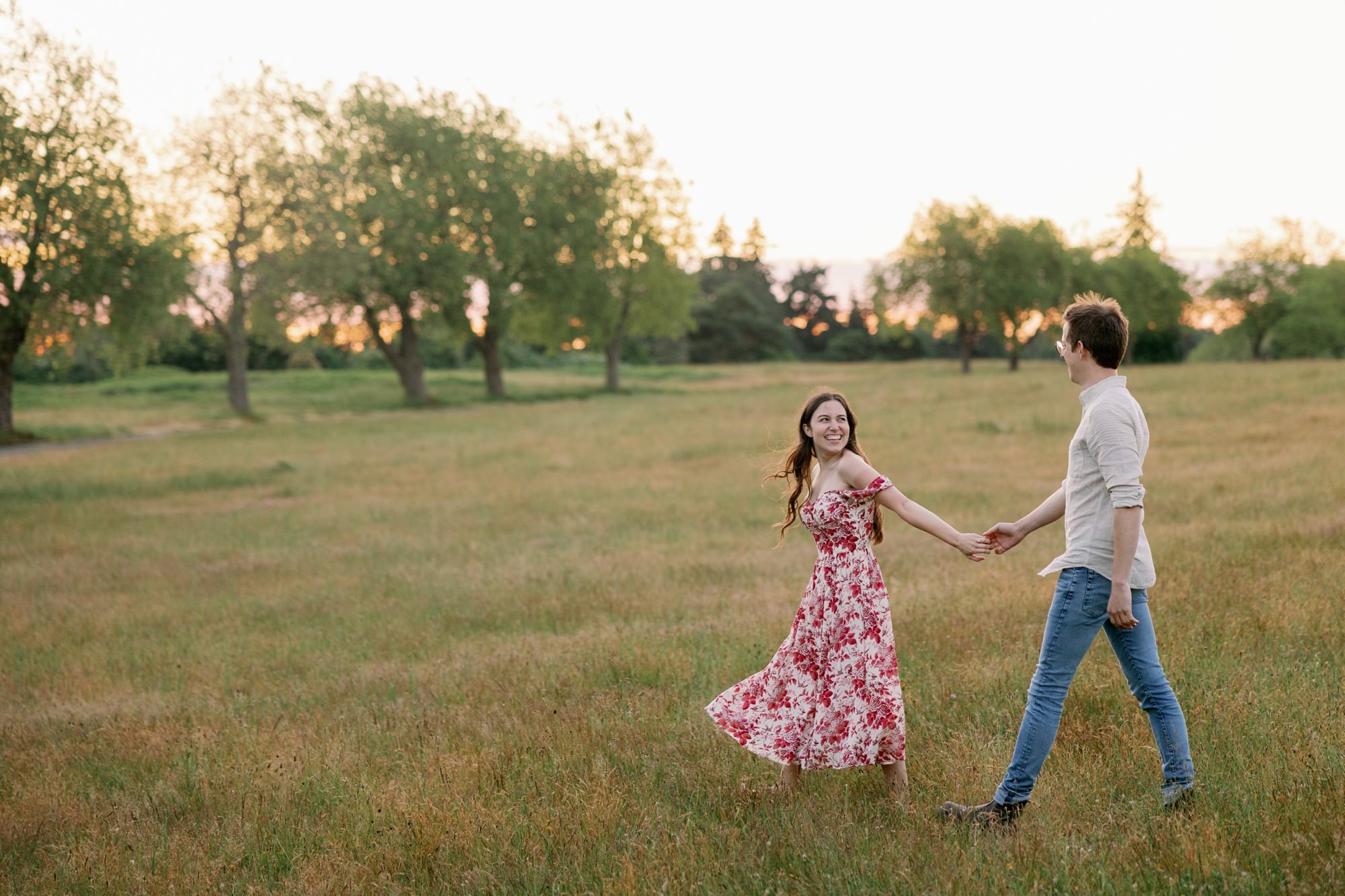 A couple holding hands and smiling in a wide-open field, with the woman in a floral dress and the man in a light shirt.