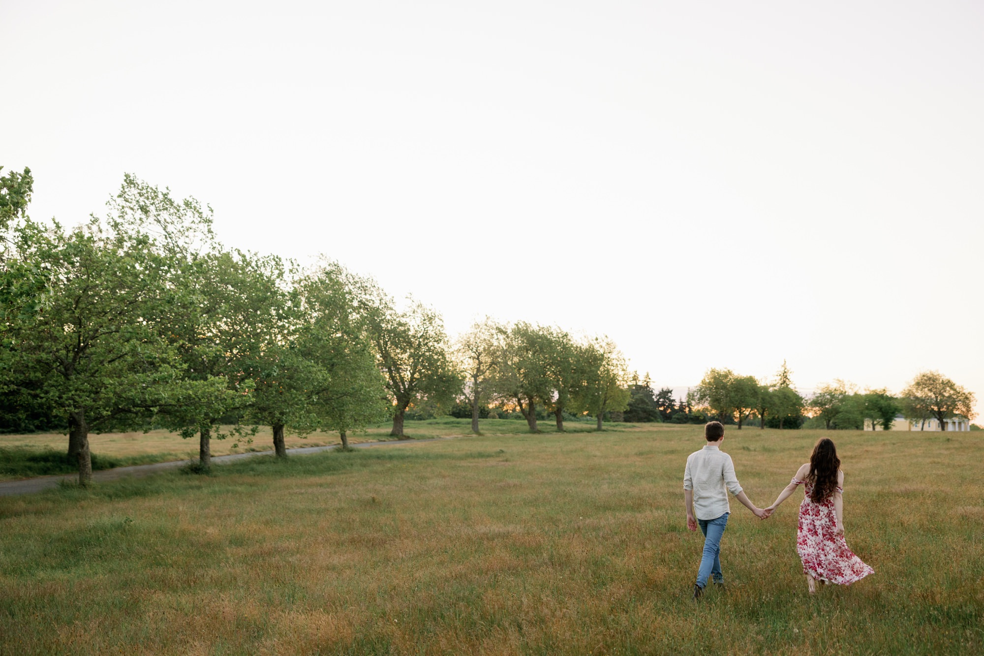 A couple holding hands and walking towards the sunset in a large open field, with the woman in a floral dress and the man in a light shirt.