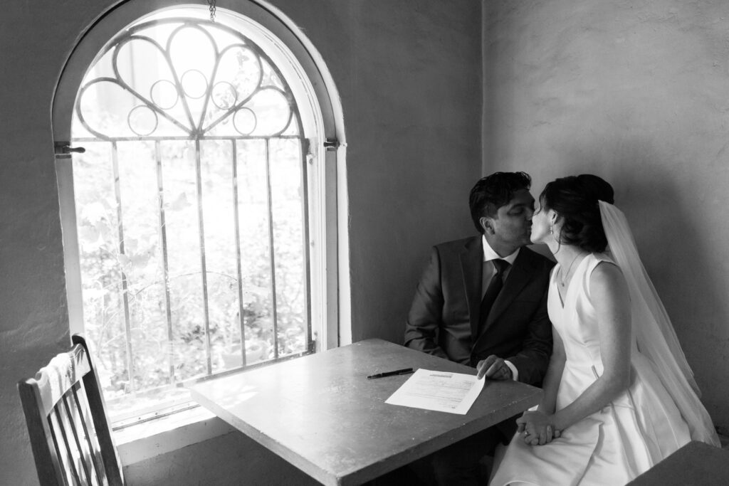Bride and groom signing their marriage certificate in a cozy room with natural light from a window.
