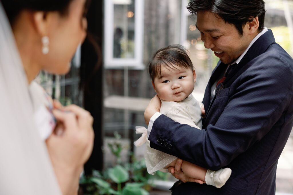 Groom holding a smiling baby while the bride looks on with a smile.