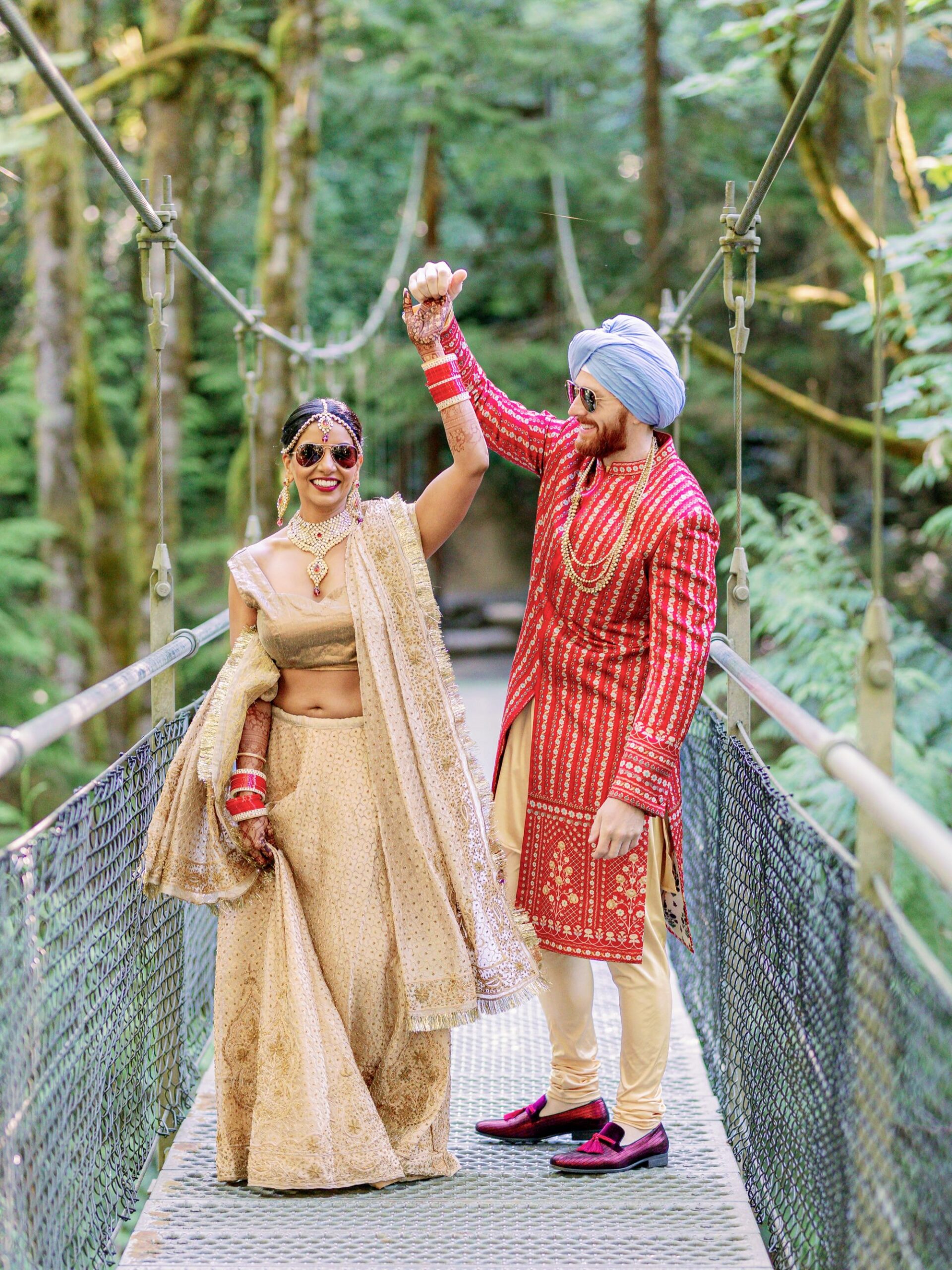 A couple dressed in traditional Indian wedding attire dancing hand-in-hand on a forest bridge.