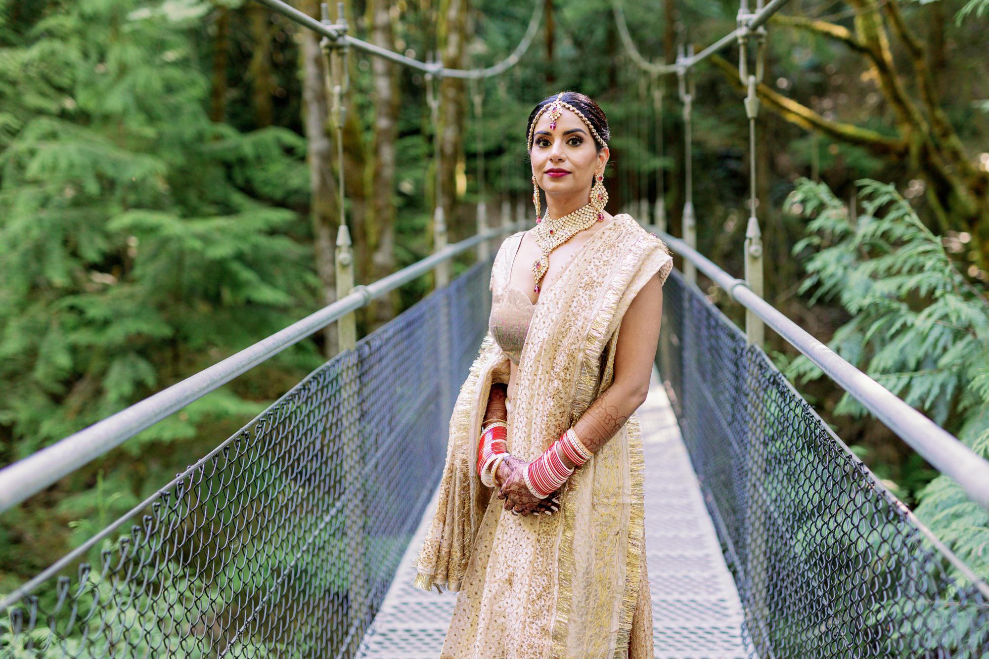 Bride in traditional Indian wedding attire standing on a forest bridge.