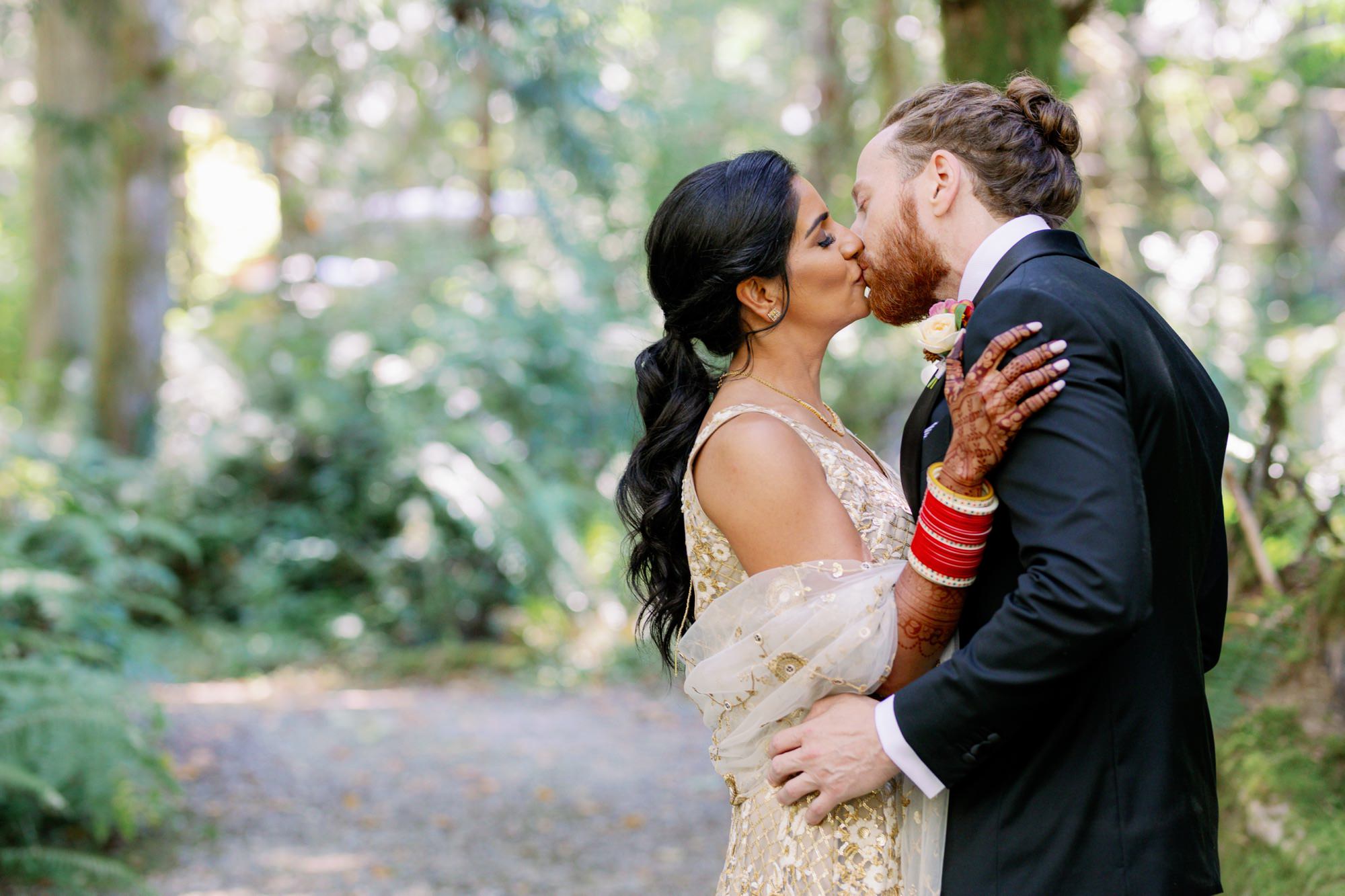 Couple embracing and kissing in a lush green forest at their IslandWood wedding.