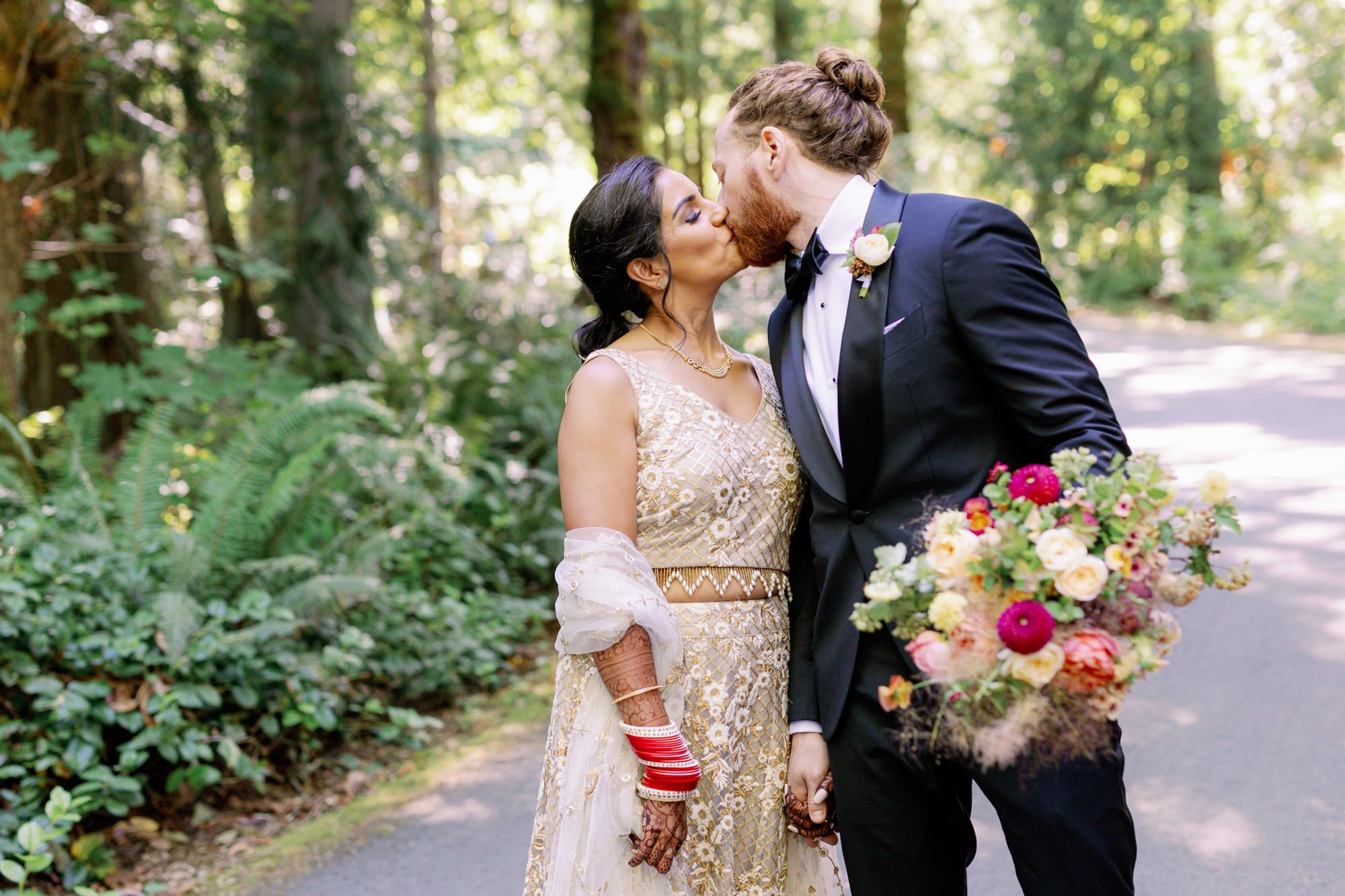 Couple sharing a kiss on a forest path, holding a vibrant bouquet.