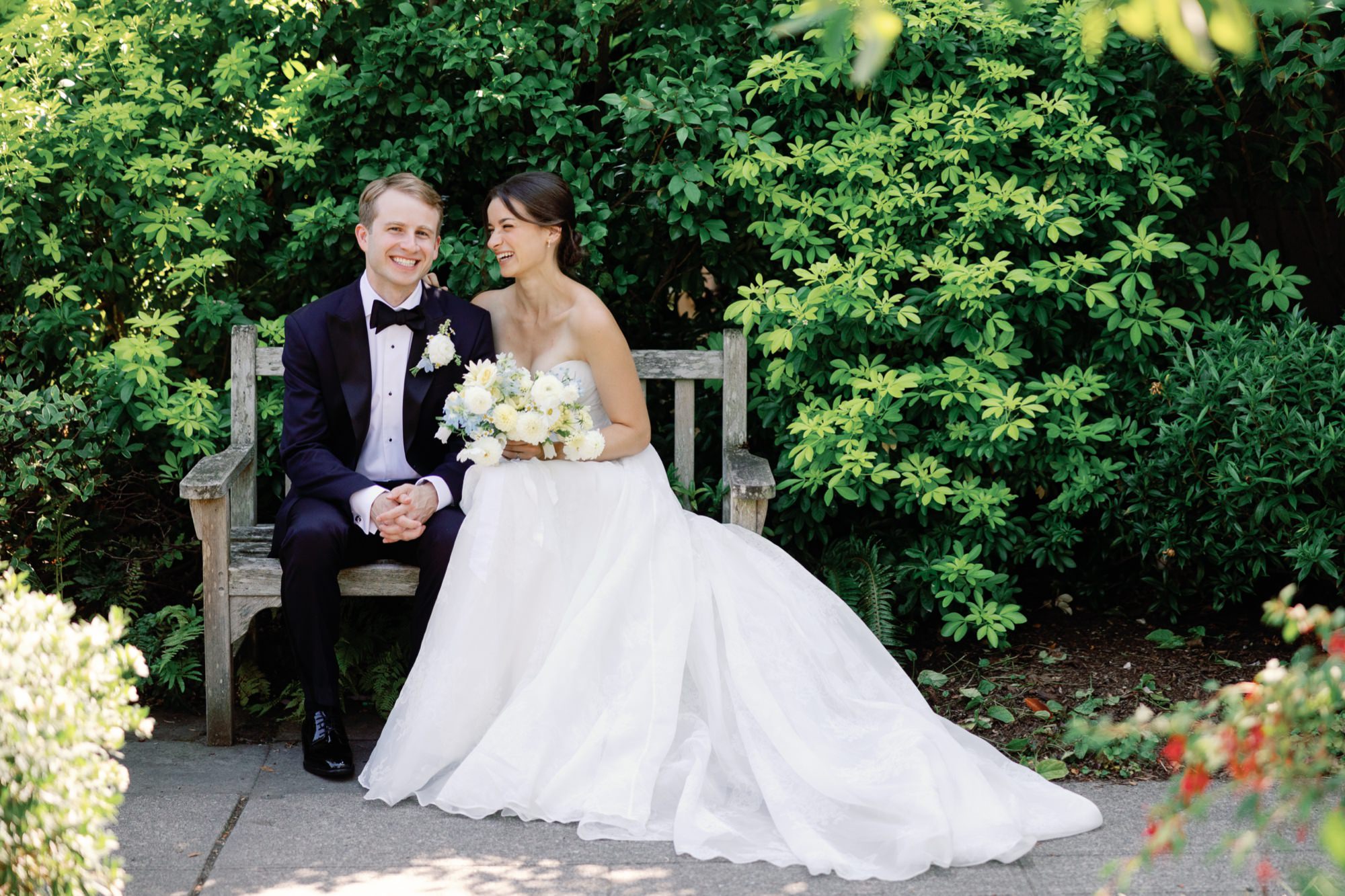 Wedding couple posing for portraits on a bench framed by green foliage.