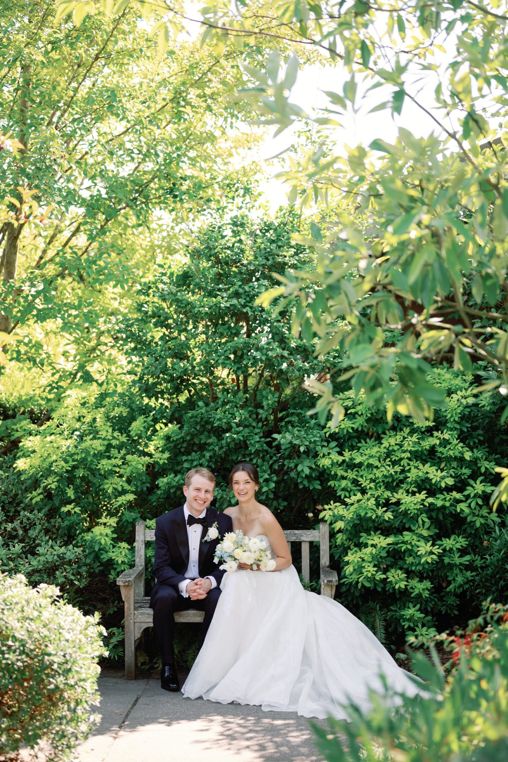 Wedding couple sits together on a bench framed by green foliage.