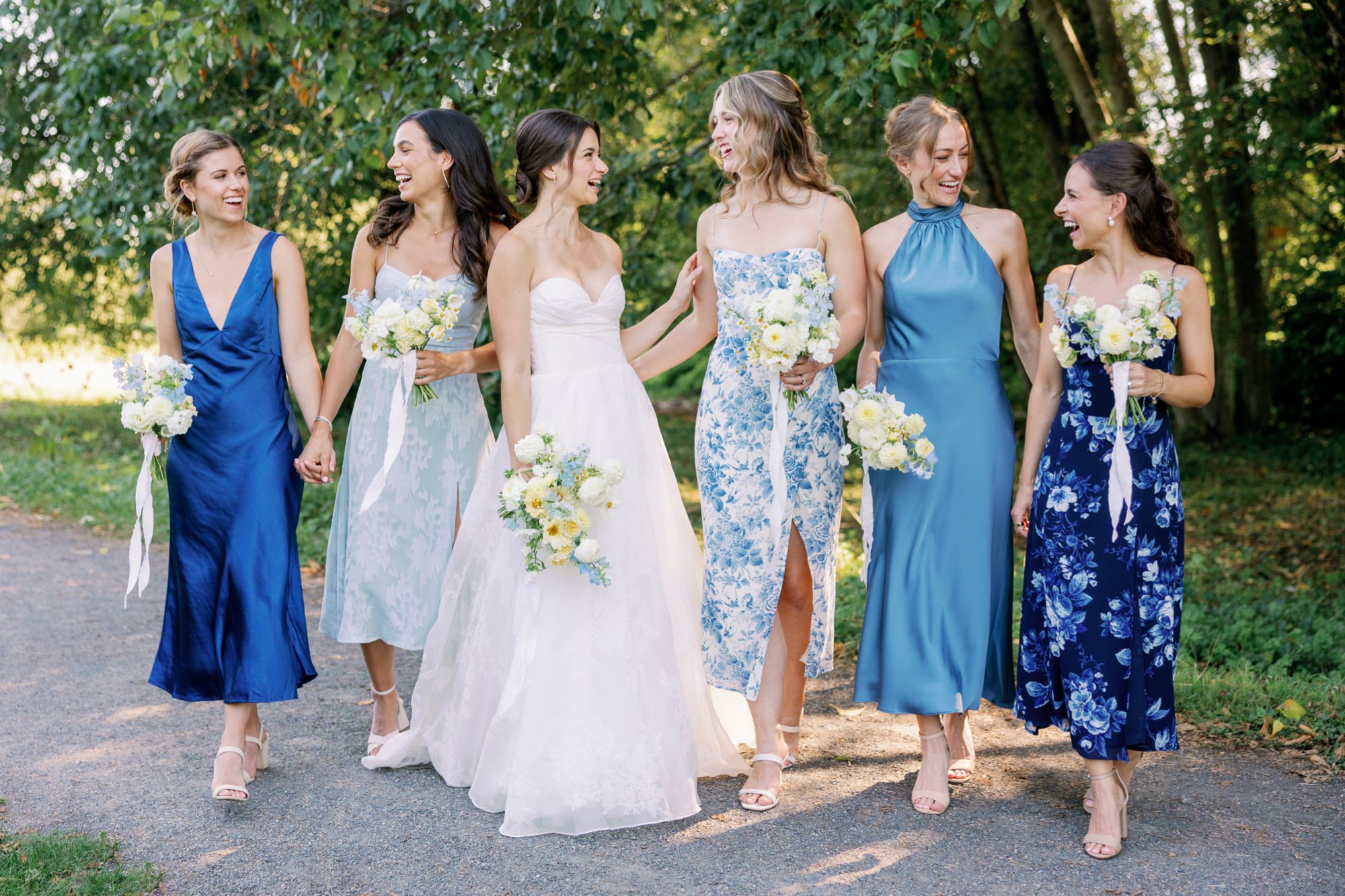 Bridesmaids and bride walking together and smiling with each other on a path with green trees in the background.