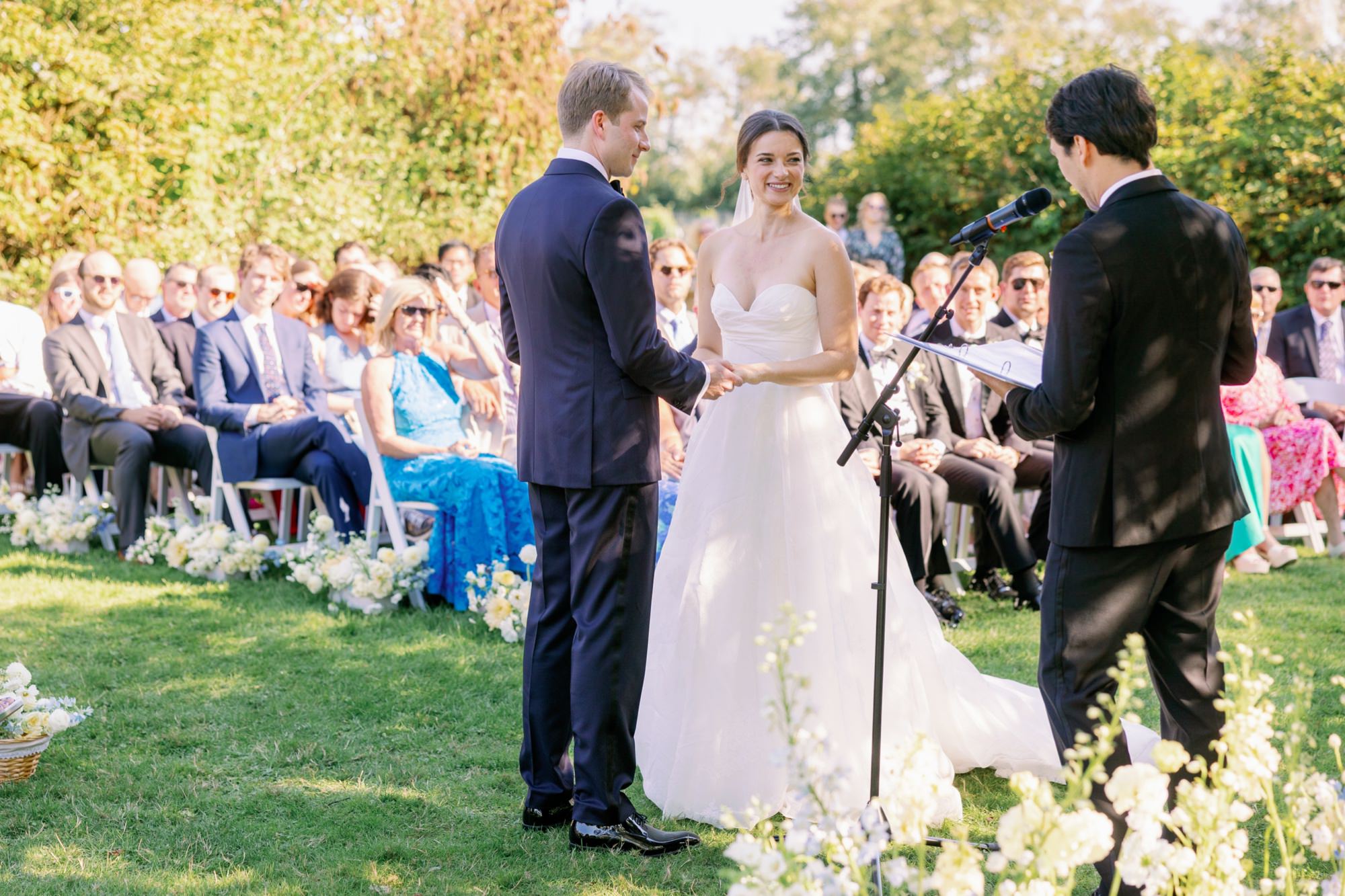 The couple looks at the officiant with guests in the background.