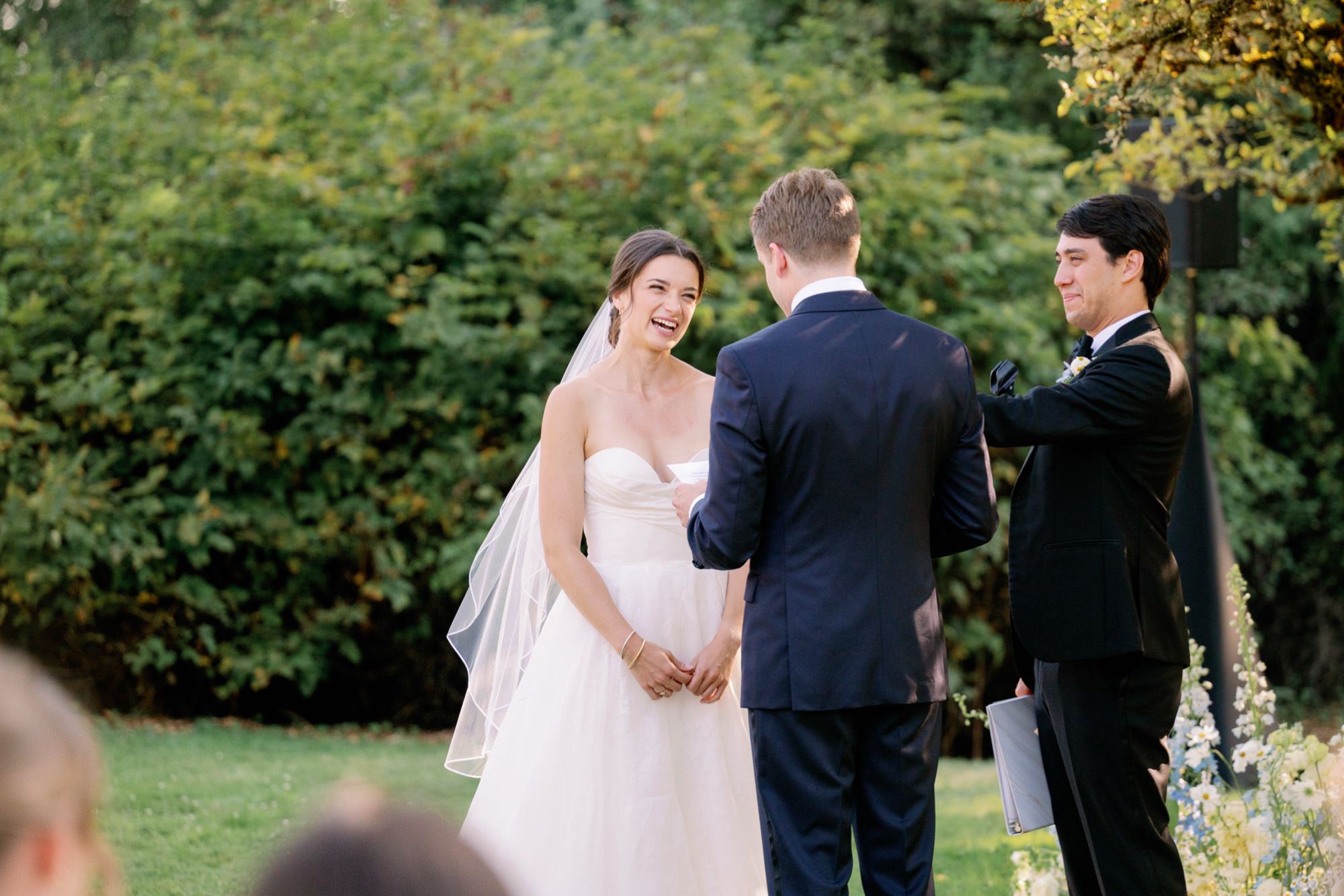The bride laughs as the groom shares his vows during an outdoor ceremony, with soft natural light and greenery surrounding the space.