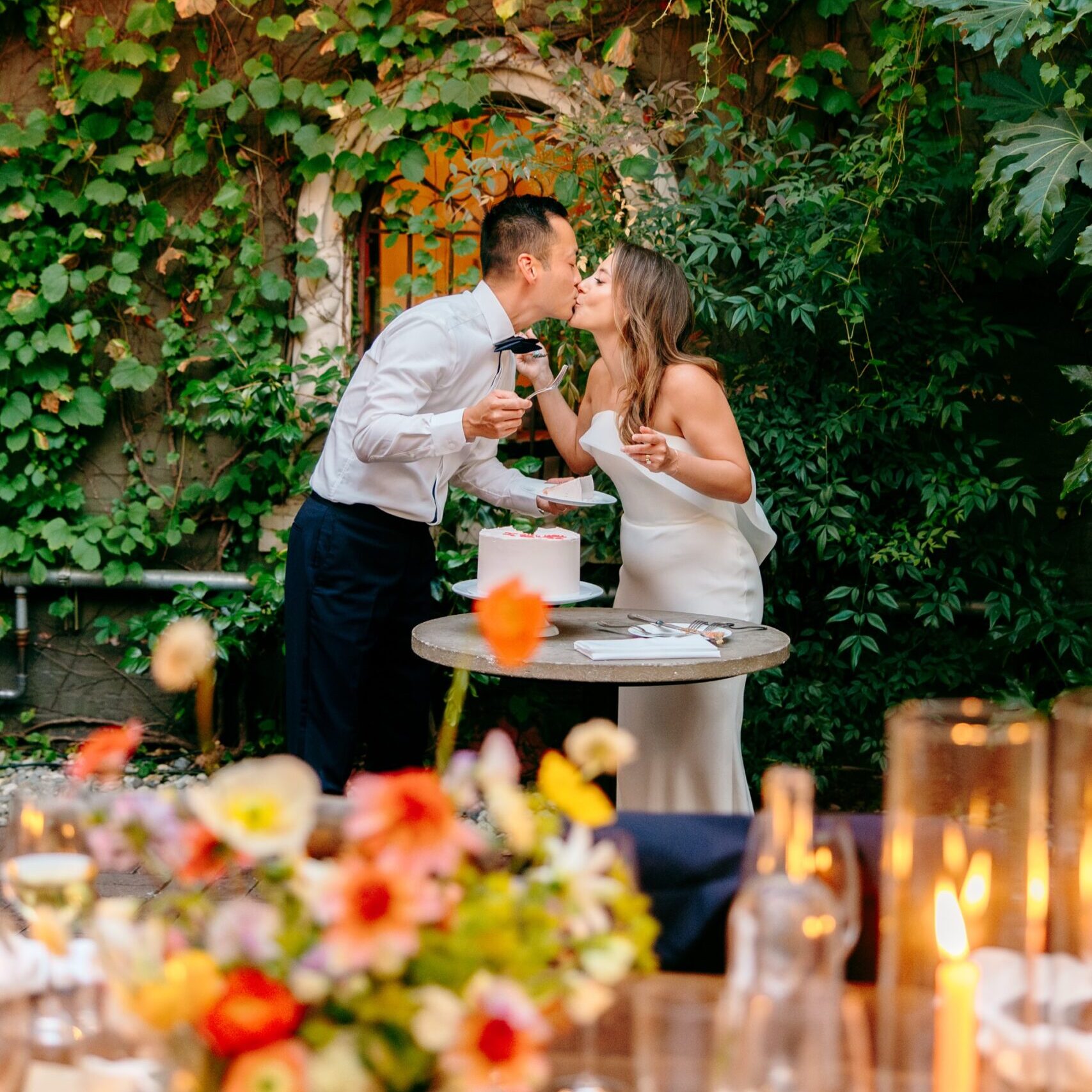 Couple kisses after cutting their cake in front of the vine-covered wall at their wedding at the Corson Building in Seattle.