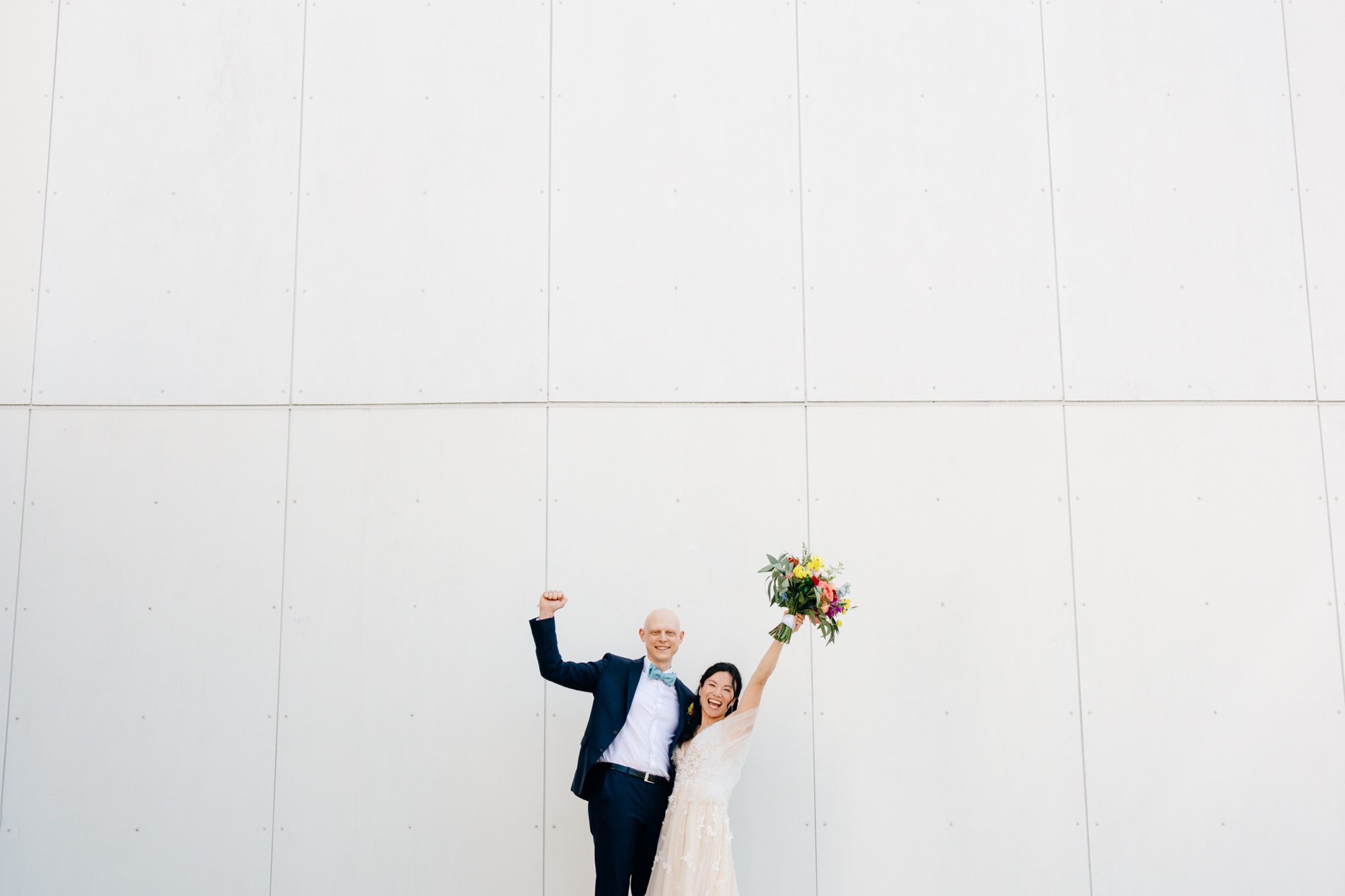 The bride and groom celebrate with arms in the air in front of a large white wall.