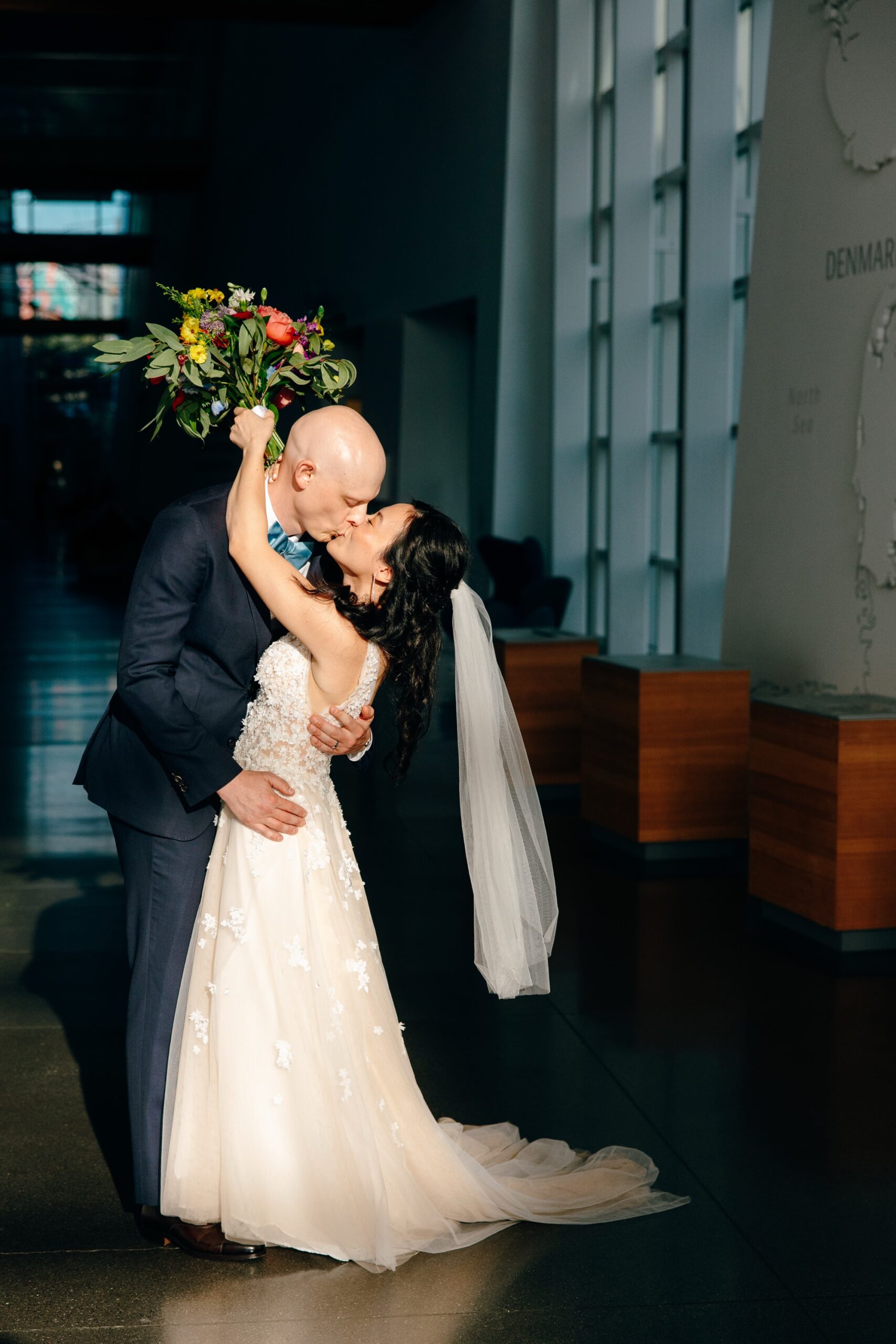 The bride holds her flowers while wrapping her arms around her husband while they kiss in Fjord Hall at National Nordic Museum just after their wedding ceremony